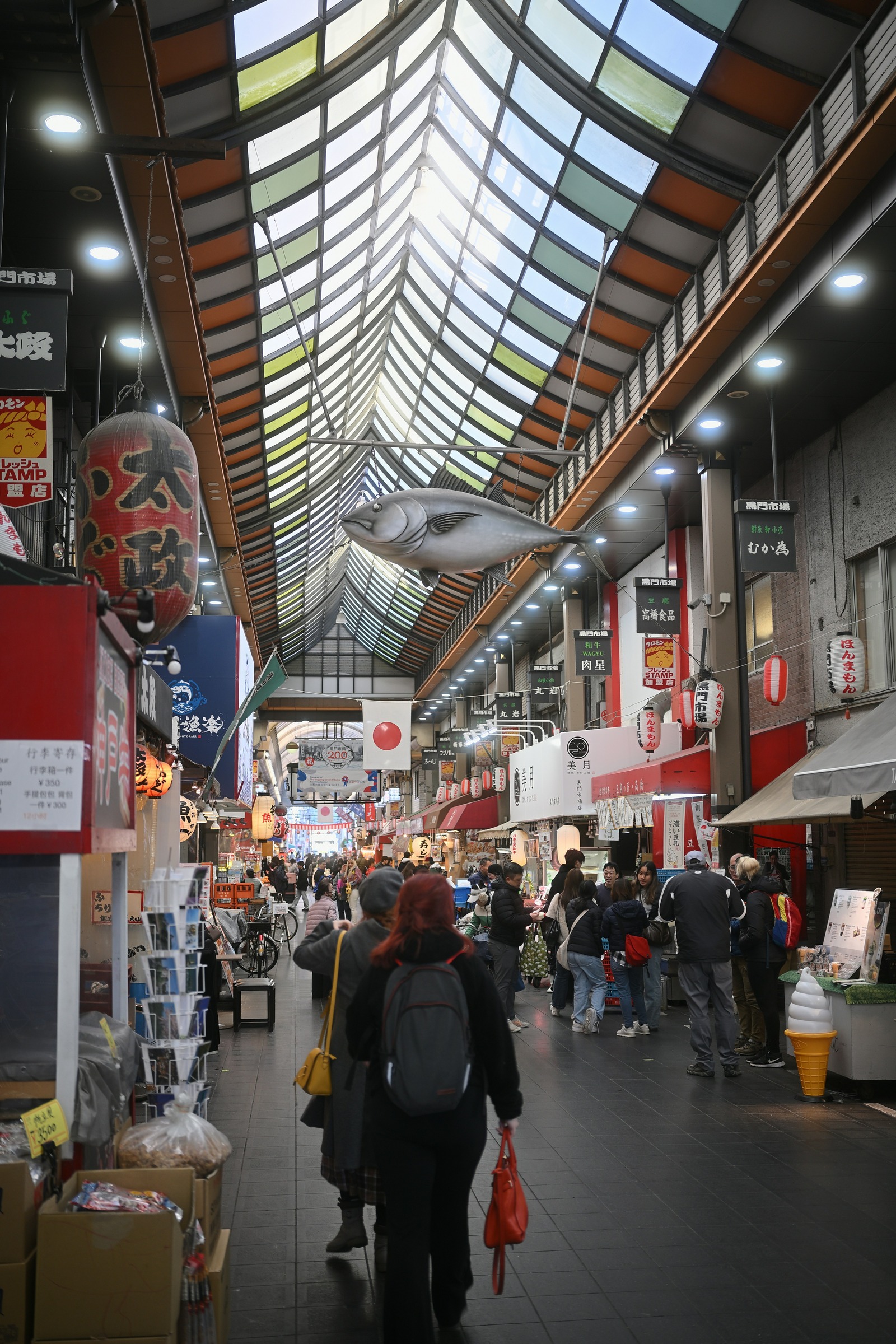 Shoppers and food stalls beneath a hanging fish sculpture in Kuromon Market