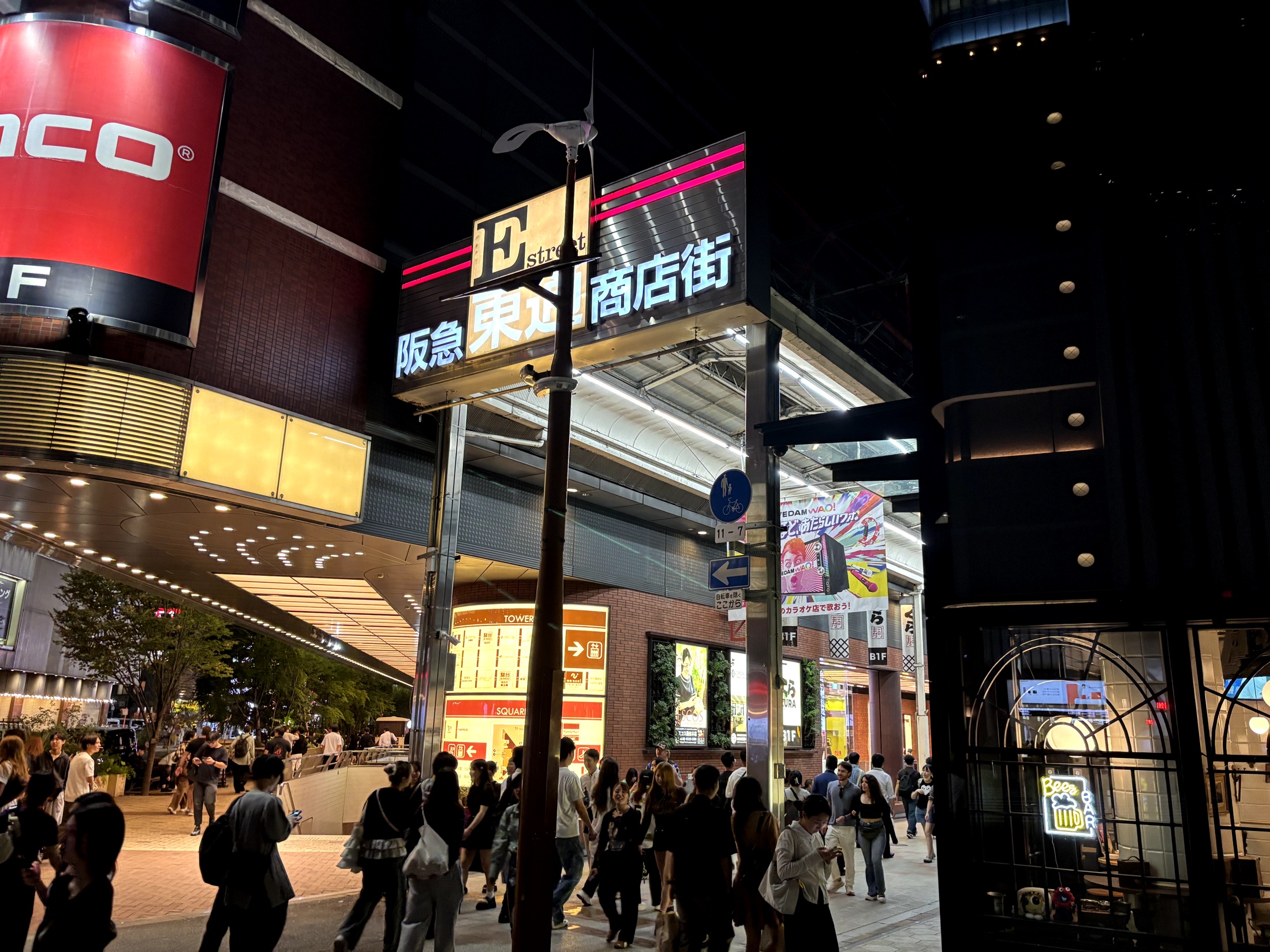 People walking past illuminated signs at the Hankyu Higashidori shopping street entrance